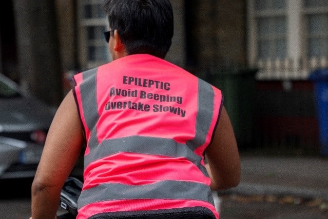 a cyclists in a high visibility bright pink cycling jacket facing away from us. The words 'Epileptic Avoid beeping overtake slowly' are written on the back of the jacket in bold text.