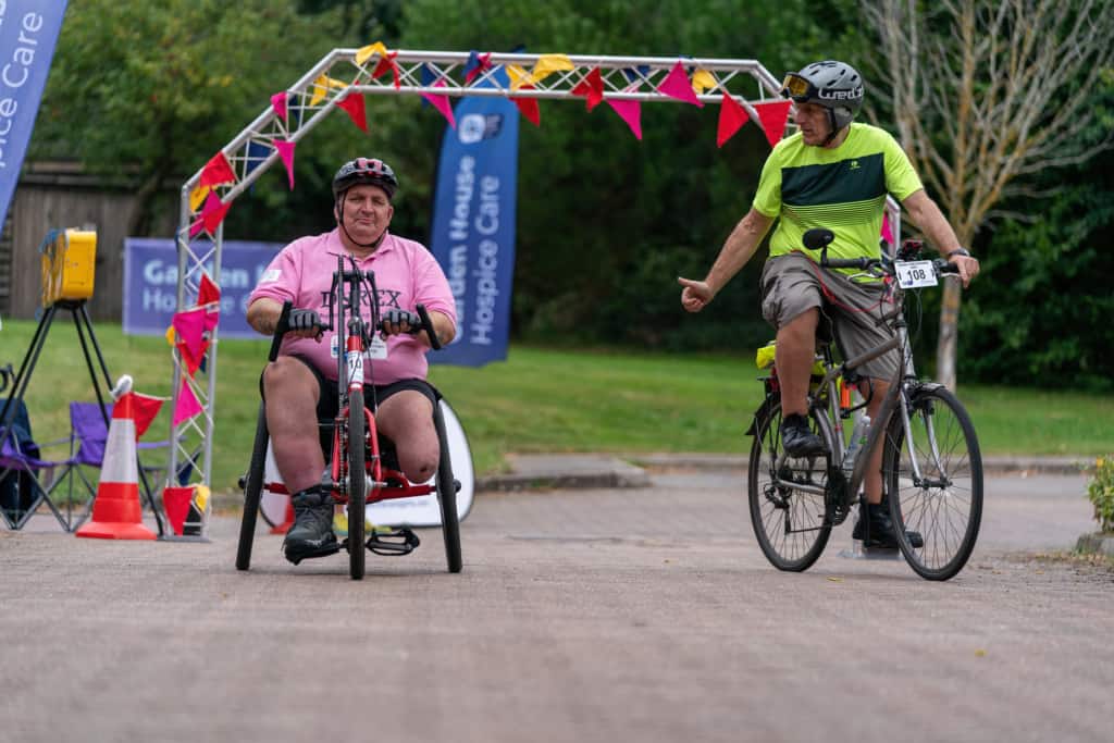 Two people cycling together, one on a two wheel bicycle and one using a handcycle, they have an archway behind them covered in bunting. The one on the bicycle has his thumb up checking and is facing toward the hand cyclist and has a caring look to his posture.  