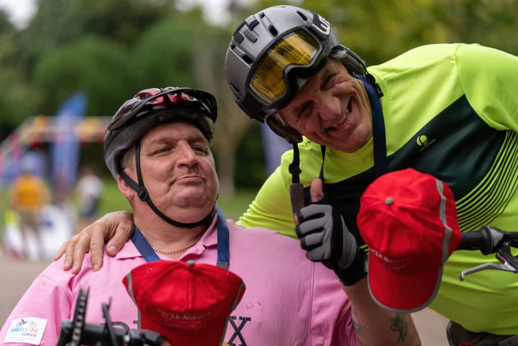 Two guys pose for the camera smiling, one in a pink polo shirt has his thumb up, they're both wearing cycling helmets. 