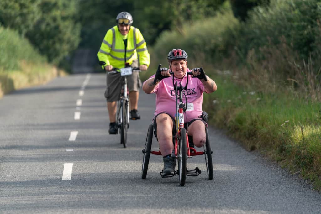 Two cyclists on a long stretch of road with grassy verges on both sides. In the foreground is a hand cyclist with one leg he is wearing a pink shirt and smiling. Following up behind him is a a smiling man using a bicycle and wearing a bright yellow high-vis jacket.