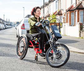 a woman using a handcycle turning towards the right of the image, the handcycle is clipped onto the front of her wheelchair  which is bright orange. 