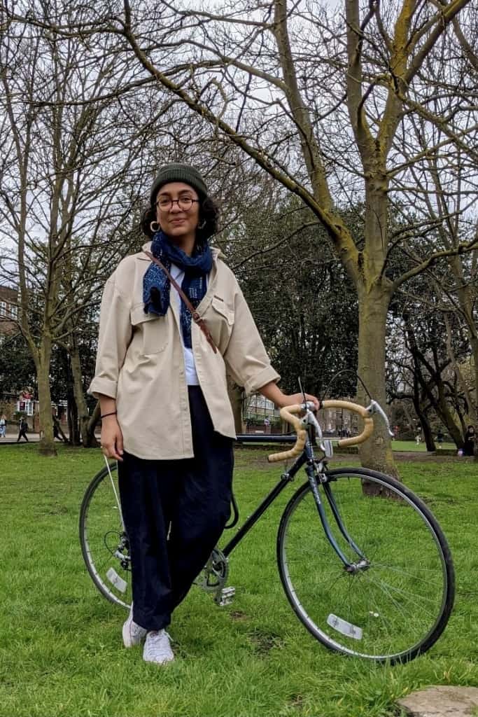 Nonki stands with her bicycle, smiling, she's wearing a woolly hat and glasses with a blue scarf, black trousers, and a bague jacket. She's on grass with trees behind her.