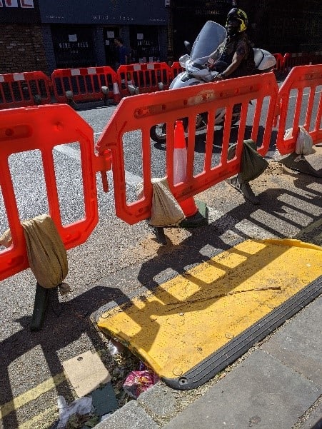 A temporary rubber curb drop leads directly into a temporary on street barrier without even the space to exit the ramp. a Moped user is driving along the road behind. 