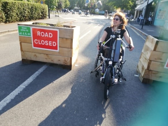 A smiling woman using a clip on Wheelchair Hand-cycle passes between on road plant boxes that are very wide apart, on one of the plant boxes the words "Road Closed" are visible in whit writing on a red background.