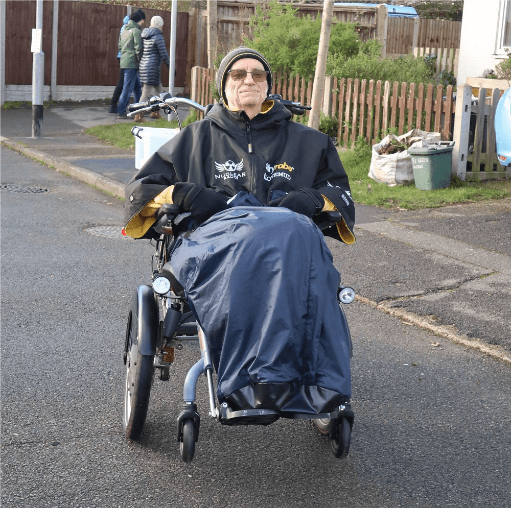 A man sits all wrapped up at the front of a Velo wheelchair cycle. you can only really see the wheelchair section in this image, with it's front casters off the floor and just the handlebars of the cycle section poking out behind the man. 