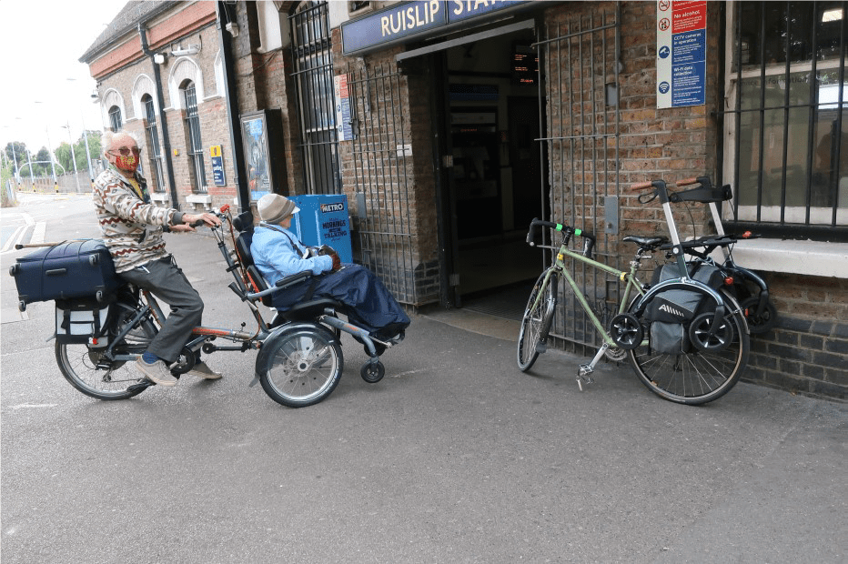 Side view of two people riding together in a Velo wheelchair cycle. They are arriving at Ruislip Station and have a suitcase attached to the back of the cycle where panniers would be. 