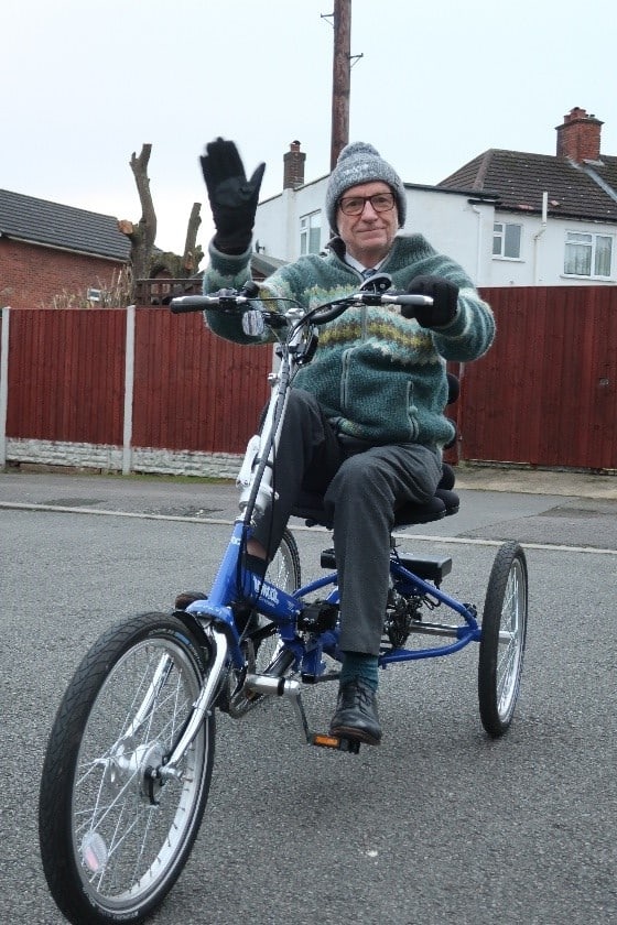 A man wrapped up warm in woolly hat and gloves waives happily sitting upon a tricycle. The trike is blue and silver and is stationary on a residential street.