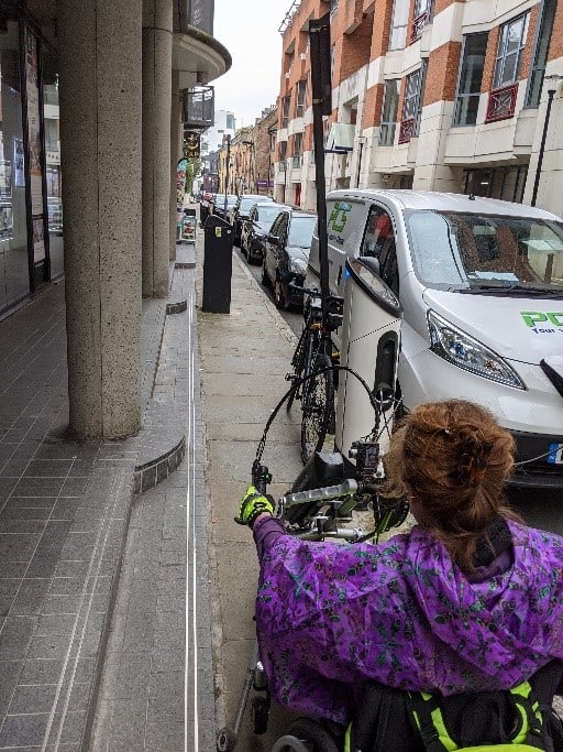 A woman dressed in purple riding a hand-cycle away from us is blocked by on street electric charging points and a street that's already too thin.