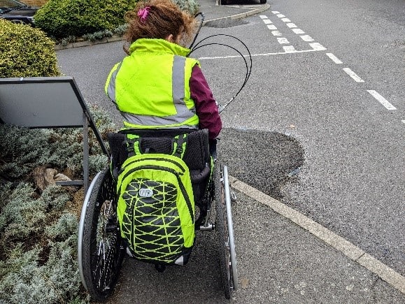 A woman with a high-visibility jacket riding a wheelchair clip on hand-cycle is stopped at the edge of a curb which has had some tarmac haphazardly piled against it to create a ramp. 