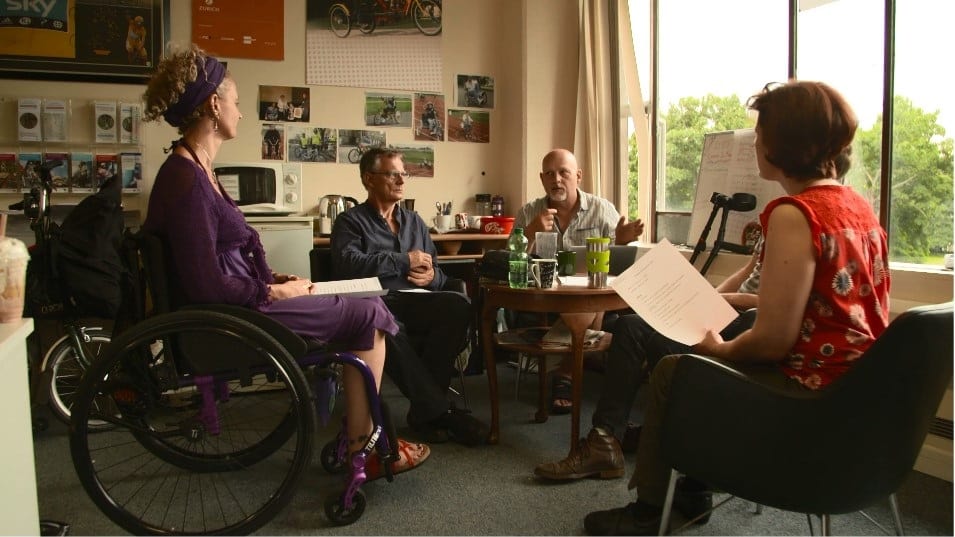 A group of people sitting around a table in discussion, papers in hand. one of them is sitting in a wheelchair, there are a pair of crutches in the background of another.
