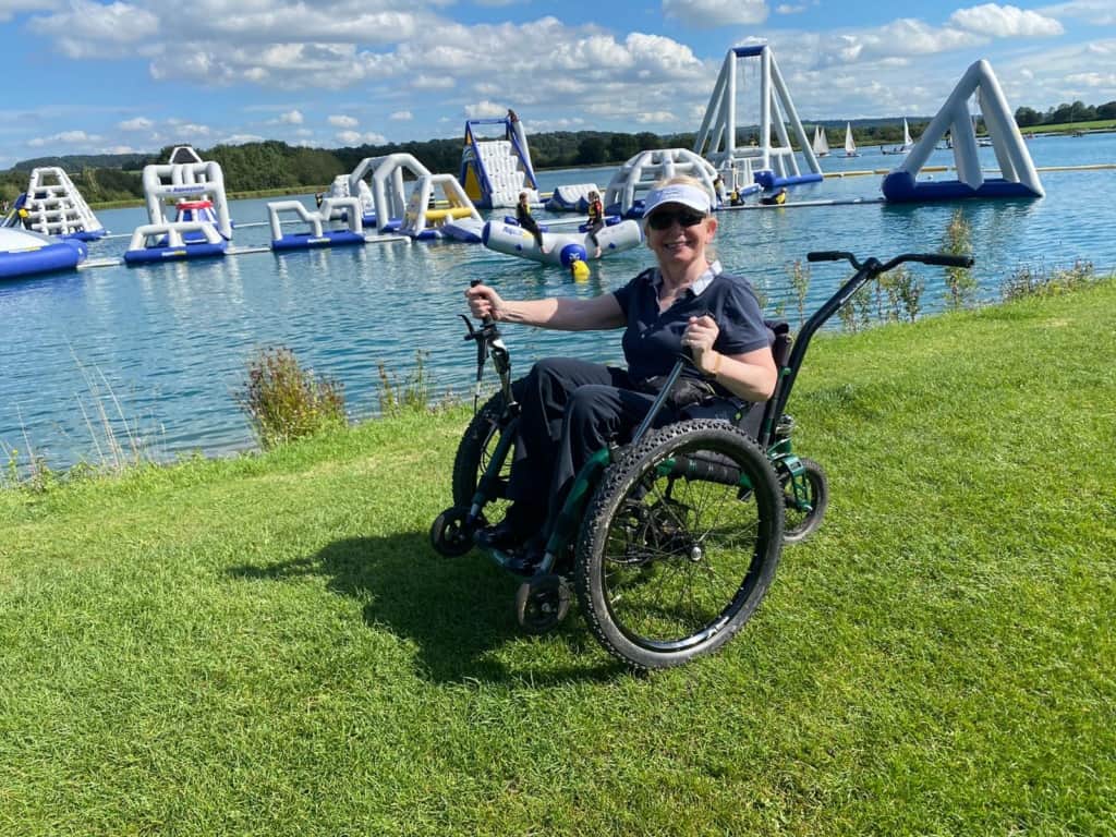 A woman sits in a mountain trike which is on grass with a lake behind her, in the lake are a number of large inflatables. The mountain trike wheelchair a 3-wheeled, all-terrain wheelchair has chain driven levers, big fat tyres and a bicycle handlebars style push handle at the back.