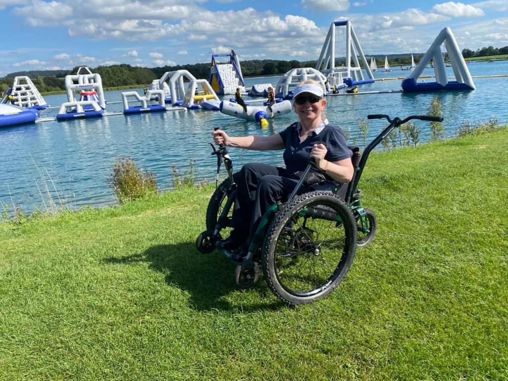 A woman sits in a mountain trike which is on grass with a lake behind her, in the lake are a number of large inflatables. The mountain trike wheelchair  a 3-wheeled, all-terrain wheelchair has chain driven levers, big fat tyres and a bicycle handlebars  style push handle at the back.