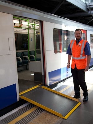 A member of train staff in high-vis vest stands next to a portable ramp, allowing access to a train. 