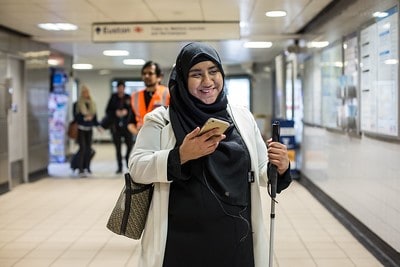 A Visually Impaired woman wearing a hijab. She is carrying a cane and looking at her smartphone while standing in a station causeway.