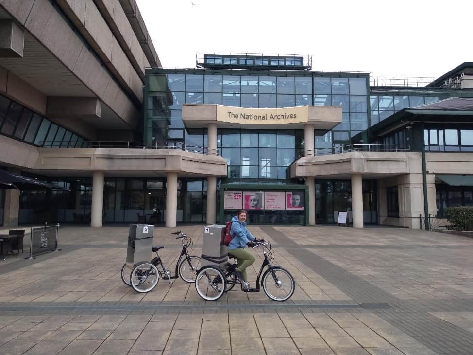 Two black trikes with silver boxes on the back in front of a glass and concrete building. A sign on the building reads 'National Archives'. A woman in a blue coat is sitting on one of the trikes.