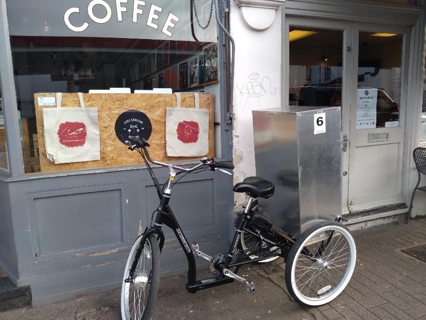 A black trike with a silver box on the back parked in front of a coffee shop.