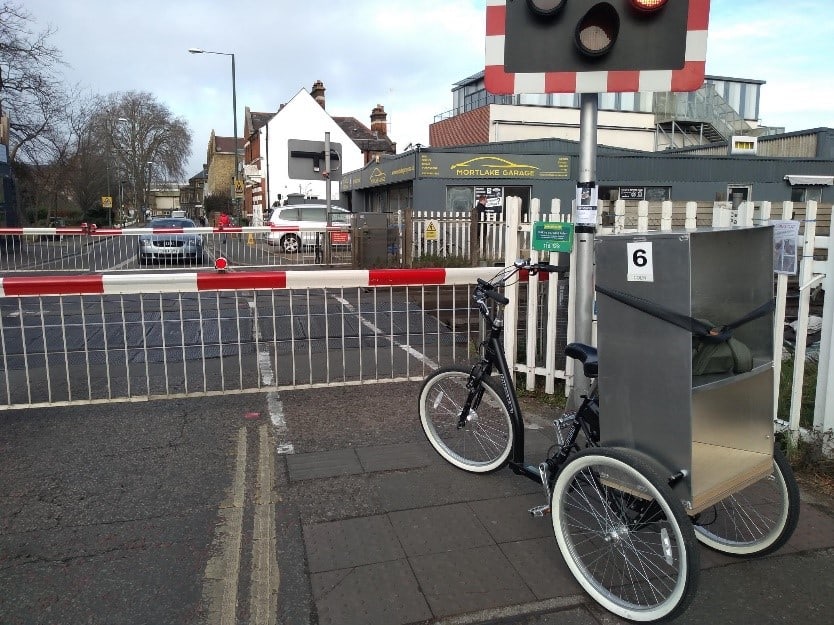 A black trike in front of a level crossing. There is a silver box on the back of the trike, with an internal shelf.
