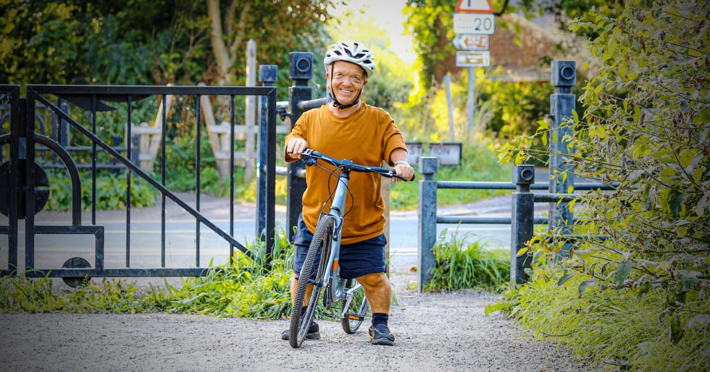 A person with disproportionate dwarfism stands with their Islabike smiling at the entry to a park. They are wearing a mustard yellow top, white cycle helmet and blue shorts.  