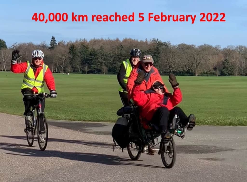 Angela and Peter cycling on a tandem cycle on a path in a park. Angela's hands are in the air. Behind them are a woman waving and a man on bicycles. 
Text across the top reads 40,000 km reached 5 February 2022.