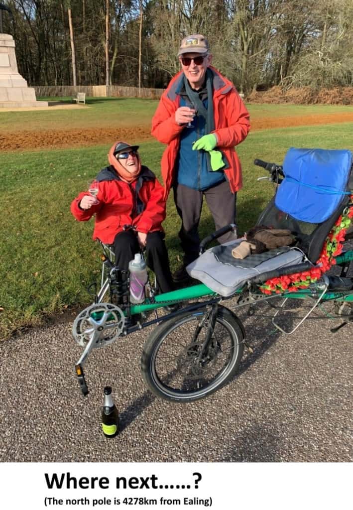 a woman sitting next to a man standing  next to a path in a green field. Both smiling holding plastic champagne glasses with a green recumbent tandem in the foreground.
Text at the bottom reads 'Where next......? (The north pole is 4278km from Ealing)'