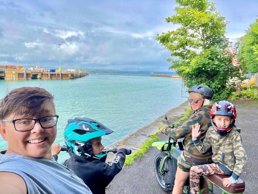 4 people, two adults and two children, sharing two cycles smiling for a selfie by the sea. Bright blue water blends with cloudy sky and bright green trees in the right hand top corner.
