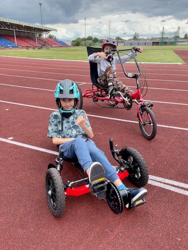 Two boys using different recumbent trikes pose for the photo while riding around a racing track. They both have BMX style closed face helmets on. 