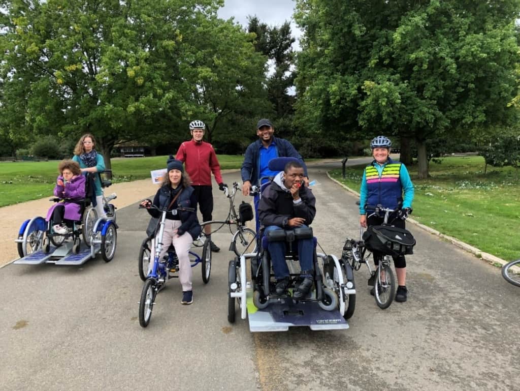 A group of individuals using non-standard cycles including Velo-plus style ride on with wheelchair cycles sit together in Dulwich park. 