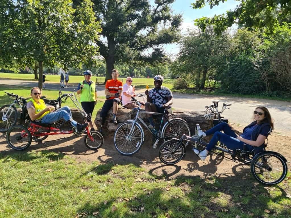 A group of cyclists looking relaxed in a park environment, they're using recumbent and non-standard cycles, a couple of them are holding ice lollies, all are smiling.