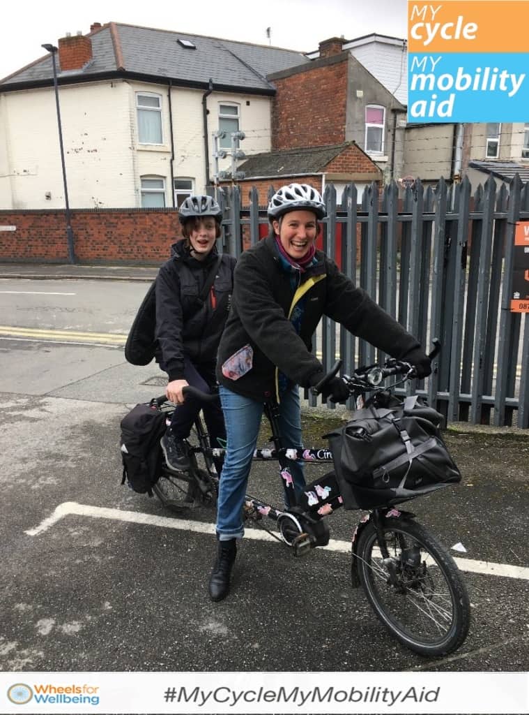Kate and her daughter smile to the camera sitting astride a tandem cycle in a suburban carpark area. the logo for #MyCycleMyMobilityAid is in the top right corner and as a banner on the bottom of the image.