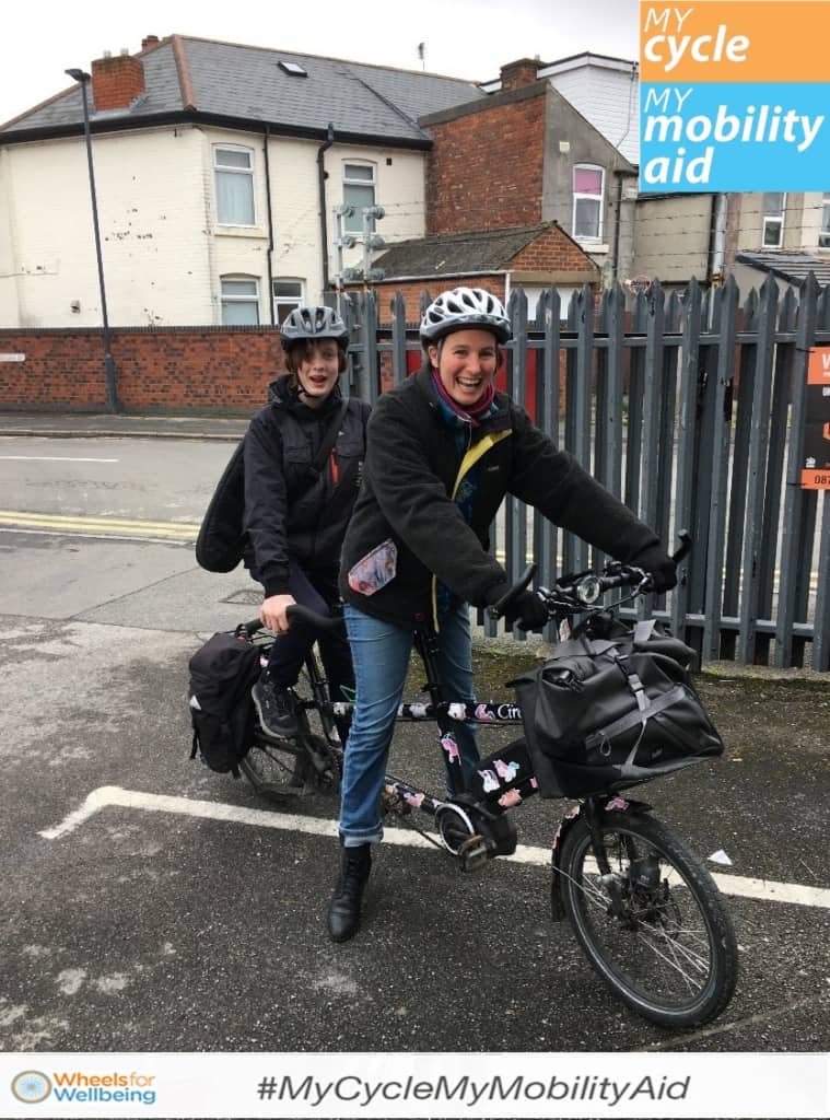 Kate and her daughter smile to the camera sitting astride a tandem cycle in a suburban carpark area. the logo for #MyCycleMyMobilityAid is in the top right corner and as a banner on the bottom of the image. 