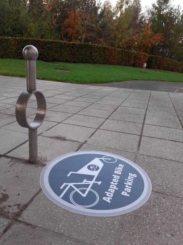 A stand design for non-standard cycles, an upright pole with a loop in the center for locking to cycles. It stands on a concrete base with a sign beneath it identifiying it as being for Adapted cycle parking. 