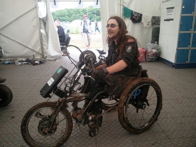 Richard Bennett aka Heavy Metal Handcyclist pictured at a festival inside a white marquee. Richard is smiling in his wheelchair with clip-on handcycle attached and muddy wheels 