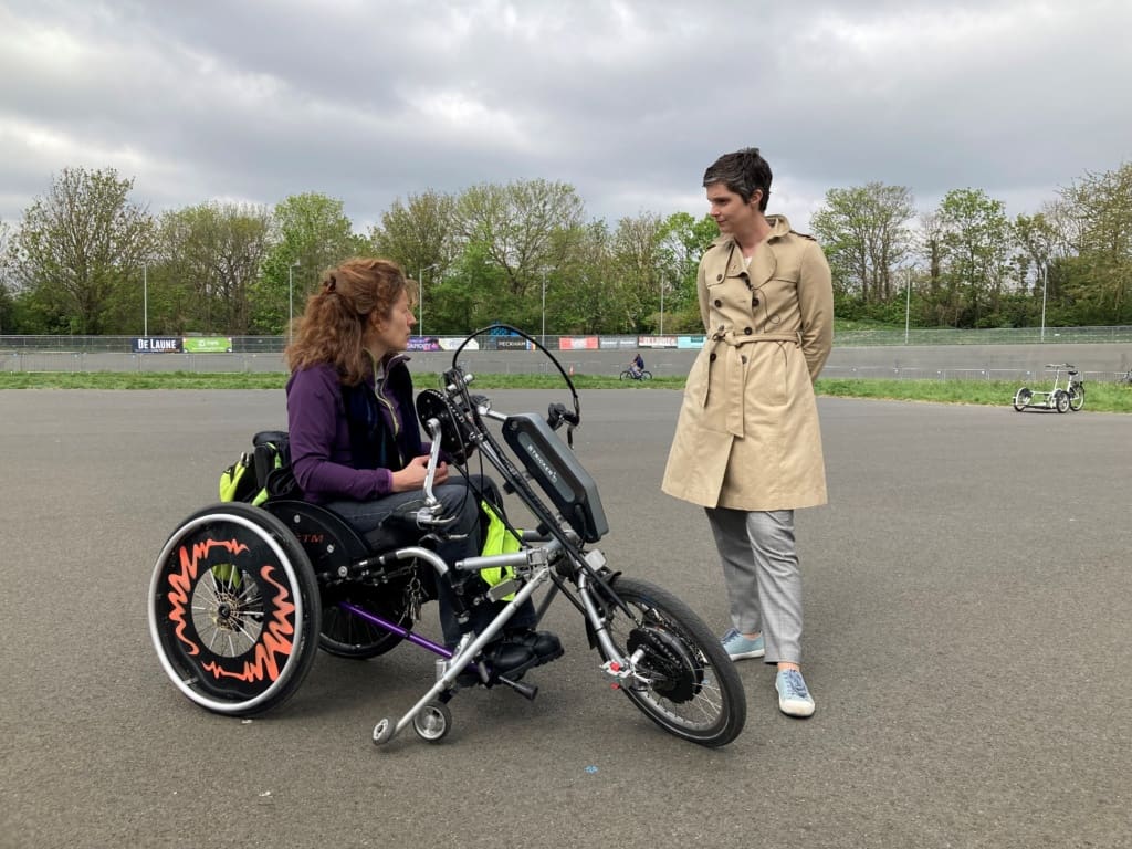 Isabelle Clement, sitting in a wheel chair with an e-assist clip-on handcycle attached talks with Chloe Smith at Herne Hill Velodrome on the inner track.