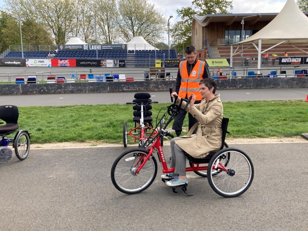 At Herne Hill Velodrome on the flat, inner tarmac track Chloe Smith is handcycling on a red upright tricycle. Ken stands behind her instructing her how to operate the cycle. 