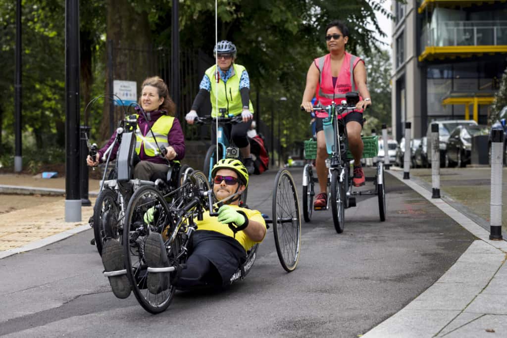 image of 3 women and 1 man riding a variety of cycles on a cycle path. They are using a standard bicycle, a tricycle, a wheelchair with handcycle attachment, and a recumbent handcycle. They are dressed in vibrant colours and hi-vis