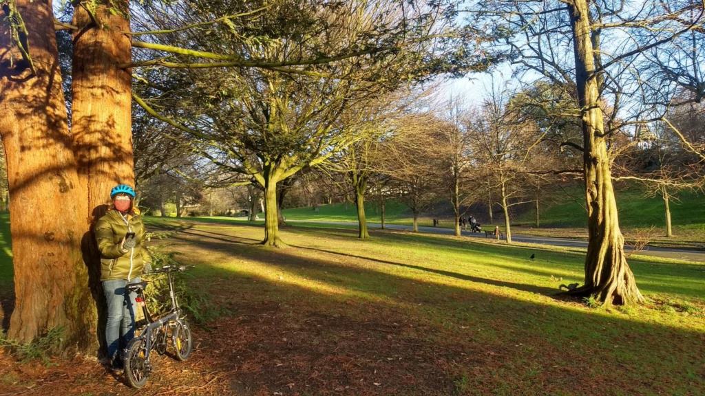 Janet is standing by a tree in a park with her bike. She is wearing a bright blue helmet and holding her phone.