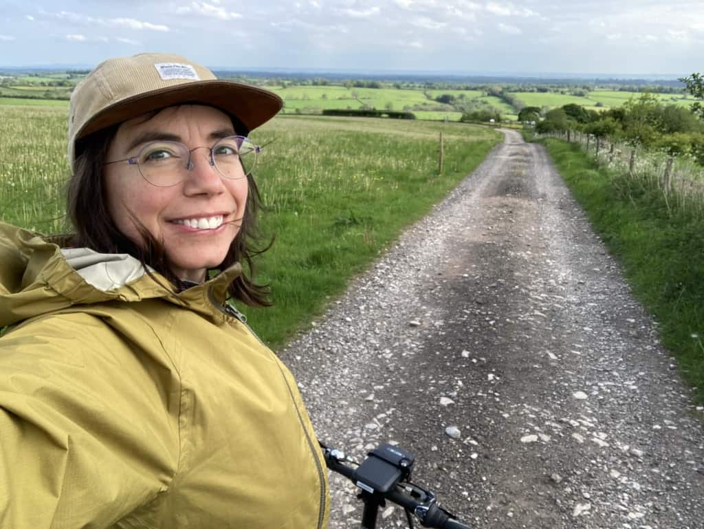 a selfie image of Janet on her bike on a country road with fields in the background