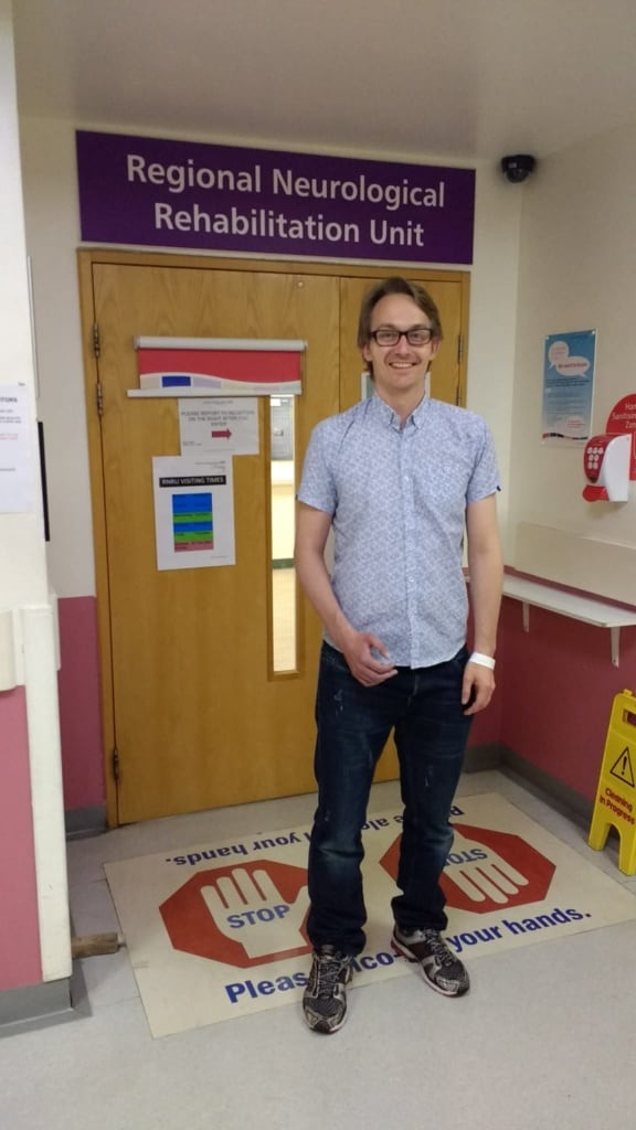 Photograph of a white man with glasses  wearing a pale blue short-sleeved shirt and dark trousers standing in front of a pale wooden door with a sign above it that reads: Regional Neurological Recovery Unit.