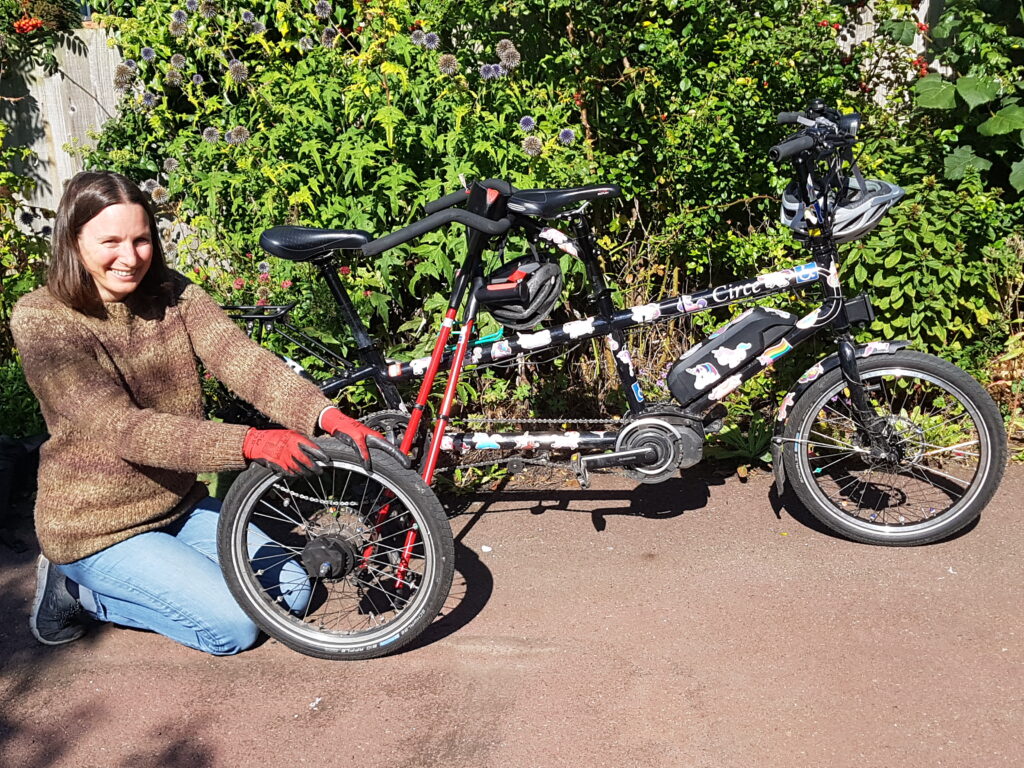 Photograph of a white woman with long brown hair kneeling beside a tandem and holding a spare wheel in her hands. The tandem is black with colourful stickers all over it and there is a pair of red crutches resting on the tandem