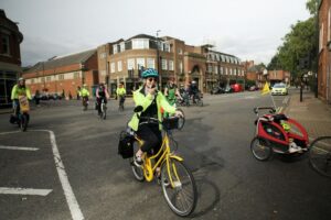 Photo shows Lucy, a white woman with a green cycle helmet, high-vis and black dungarees riding a bright yellow upright bike on a road. There are lots of other people on cycles around. Lucy is holding a microphone to her mouth and talking as she rides.