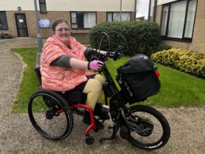 A white woman in a pink jacket is smiling at the camera. She is sitting in a wheelchair with a red frame and using a black clip-on handcycle with a chunky tyre and big black bag attached to ride up a path.