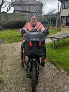 A white woman in a pink jacket is riding a handcycle down a path straight towards the camera.