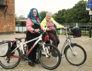 Photo shows two white women with glasses smiling at the camera. One woman has blue hair and is standing holding a red and white bike. Slightly behind her is another woman with grey hair wearing a high-vis coat and sitting on a silver e-trike. They are on a stone slab surface near a brick building with a river behind them. There is a cycle path sign in the background.