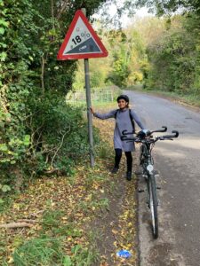 A woman in a grey dress and black leggings is standing holding the back of a tandem with one hand and a road sign showing 18% incline with the other.