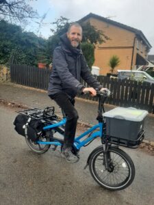 A man with a short beard riding a blue Tern HSD cargo cycle is stationary on a road smiling at the camera. There is a plastic box on the front of the cycle and a pannier on the back.