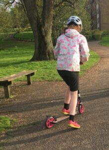 A person is scooting away from the camera wearing black shorts, pink shoes, a grey cycle helmet and a coat covered in flamingos.