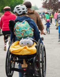 Two people are cycling away from the camera on a tandem. There is a wheelchair mounted on the back of the tandem and a yellow rain cover on the wheelchair cushion. There is a cuddly grey cat on the cushion, too.