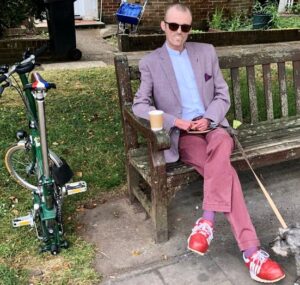 A white man sitting on a bench wearing casual jacket, shirt and sunglasses has a part-folded racing green Brompton bicycle beside him.