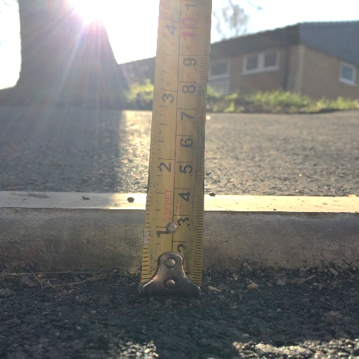 A yellow metal ruler held against a kerb edge showing approx 3cm step up onto a sloping asphalt pavement. There is a steep slope uphill to grass and a community centre building in the background.