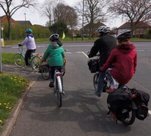 Photo shows a road joining a traffic-free cycle path. There is no barrier between the road and cycle path. A group including a woman towing a child trailer, a child riding a single bike and an adult and child on a tandem with panniers are turning onto the cycle path.